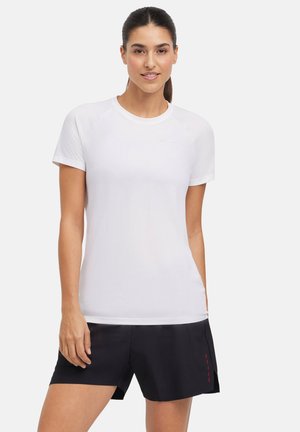 T-shirt de sport blanc à manches courtes avec une texture lisse, comportant des manches raglan. Porté avec un short noir avec des accents de couture rouges.
