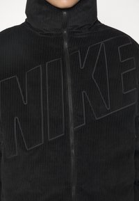 Svart kordfloret zip-jacka med hög krage och stor präglad "NIKE"-logotyp på framsidan. Texturerat tyg med vertikala linjer.