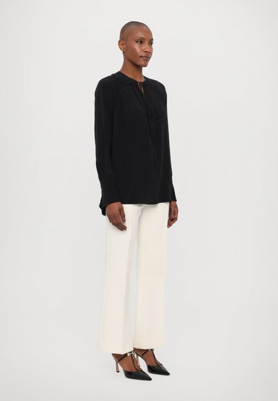 Blusa nera a maniche lunghe con scollatura rotonda e dettagli arricciati, abbinata a pantaloni bianchi a campana e scarpe nere con punta affusolata.