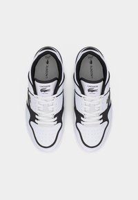 Λευκά και μαύρα αθλητικά παπούτσια Lacoste με κορδόνια, ορατό λογότυπο της μάρκας στη γλώσσα και την πλευρά, απεικονισμένα από πάνω σε λευκό φόντο.