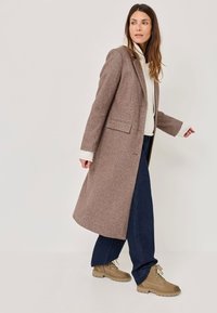 Ruskea villacoata, jossa on pitkä ja suora muotoilu, varustettuna viistolla kauluksella ja sivutaskuilla, yhdistettynä sinisiin leveälahkeisiin farkkuihin ja ruskeisiin nauhakenkiin.