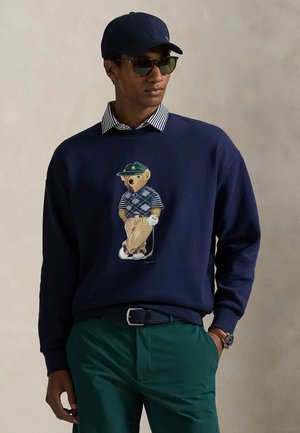 Μπλε σκούρο φούτερ με σχέδιο αρκούδας που φοράει πουλόβερ με σχέδιο, πράσινο καπέλο, γυαλιά ηλίου και πράσινο παντελόνι, με ριγωτό γιακά ως λεπτομέρεια.