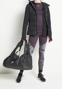 Persona con chaqueta acolchada negra, ropa deportiva púrpura de Adidas, zapatillas negras de Adidas y sosteniendo una bolsa de gimnasio negra de Adidas contra un fondo blanco liso.