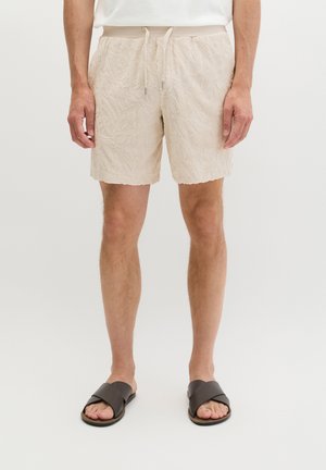 Mann trägt beige strukturierte Shorts mit Kordelzug und schwarze Sandalen mit Kreuzriemen, steht vor einem schlichten hellen Hintergrund.
