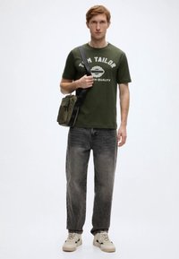 Λαδί βαμβακερό t-shirt με λευκή εκτύπωση, συνδυασμένο με γκρι τζιν με φθορές και μπεζ αθλητικά παπούτσια. Μικρή ώμου τσάντα σε λαδί χρώμα φορεμένη χιαστί στο σώμα.