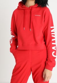 Felpa corta rossa in misto cotone, con cappuccio regolabile con coulisse e logo "CALVIN KLEIN" stampato in bianco su entrambe le maniche.