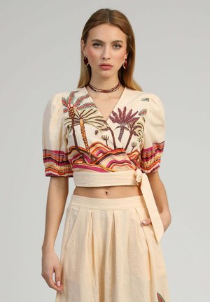 Femme portant un crop top beige cache-cœur avec imprimé coloré de palmiers et une jupe plissée taille haute assortie, posant devant un fond uni.
