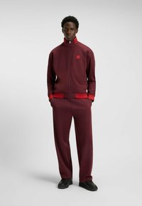 Tuta marrone con strisce rosse sulle maniche e sui pantaloni. Giacca con zip presenta un colletto alto e un logo. Vestibilità comoda e tessuto morbido.