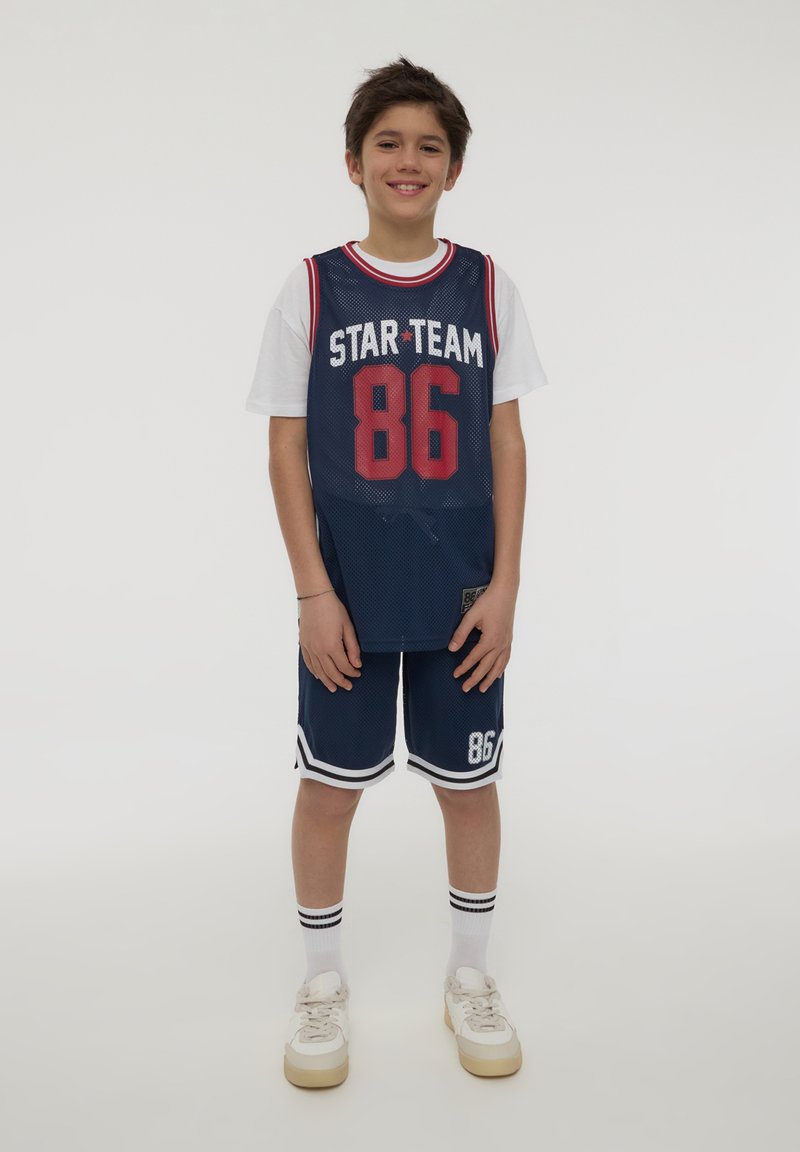 Maglia da basket blu navy con accenti rossi, tessuto a rete, canotta bianca sotto, pantaloni corti abbinati con strisce bianche e sneakers crema.