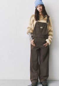 Braune Denim-Latzhosen mit weitem Bein, Vordertasche und verstellbaren Trägern, kombiniert mit einem gestreiften gelben Pullover und einer blauen Mütze.