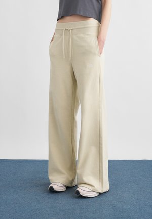 Persoon die beige wijde joggingbroek met trekkoord in de taille en lichtgrijze sneakers draagt, staand op een blauw tapijt tegen een witte muur.