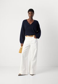 Marineblauwe v-hals trui, witte wijde broek, gele riem en bijpassende handtas. Gelaagd stof op de trui, beige en zwarte puntige schoenen.