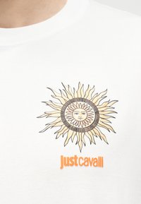 Εικονογράφηση ήλιου με πρόσωπο πάνω από το πορτοκαλί κείμενο "just cavalli" στη λαιμόκοψη λευκού T-shirt.