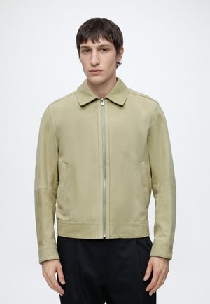 Mann mit kurzen braunen Haaren trägt eine hellbeige Jacke mit Reißverschluss und Kragen sowie schwarze Hose, steht vor einem schlichten weißen Hintergrund.