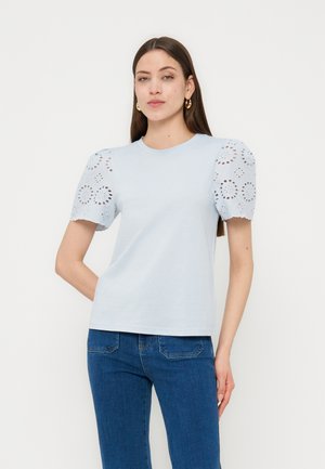 Femme portant un haut bleu clair à manches bouffantes avec des détails en broderie anglaise et un jean bleu, se tenant devant un fond blanc uni.