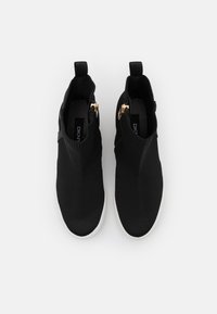 Černé pletené kotníkové boty s hladkou texturou, zdobené zipem a kontrastními bílými gumovými podrážkami. Pohled zeshora ukazuje elegantní design.
