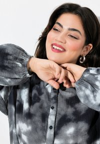 Grijze tie-dye shirt met zwart-witte patronen, lange poefmouwen, knoopsluiting aan de voorkant en subtiele textuur. Zilveren ringoorbellen zichtbaar.