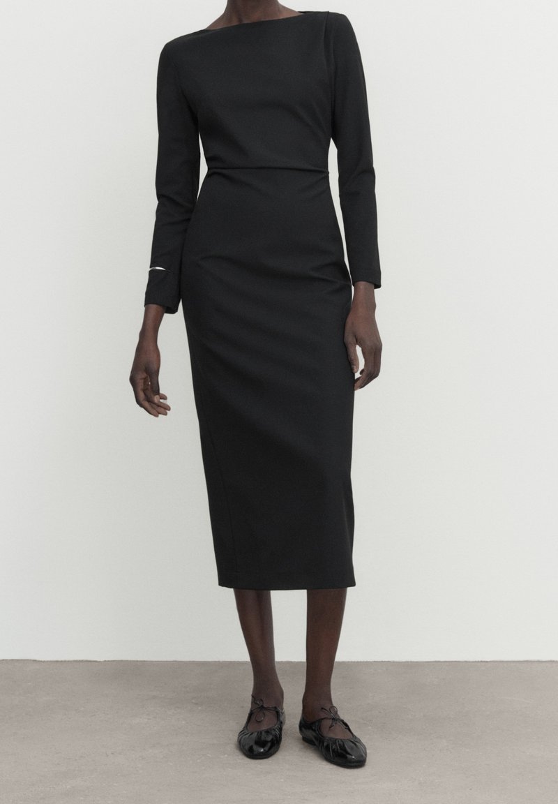 Femme portant une robe midi noire moulante à manches longues avec un col bateau et des ballerines noires, debout sur un sol gris.