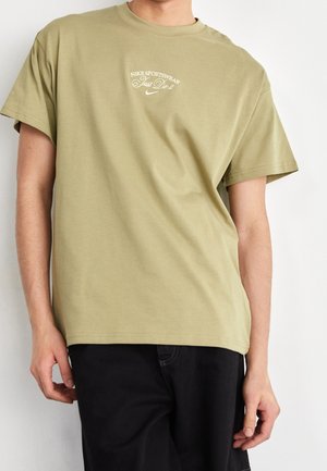 Mann trägt ein olivgrünes Nike Sportswear T-Shirt mit weißem "Just Do It" Text und schwarzen Hosen vor weißem Hintergrund.