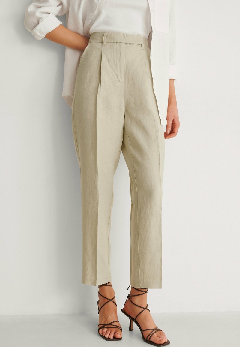 Pantalons en lin beige plissés avec une taille haute et des jambes fuselées, associés à des sandales à talons bruns à lanières. Texture lisse et design ajusté.