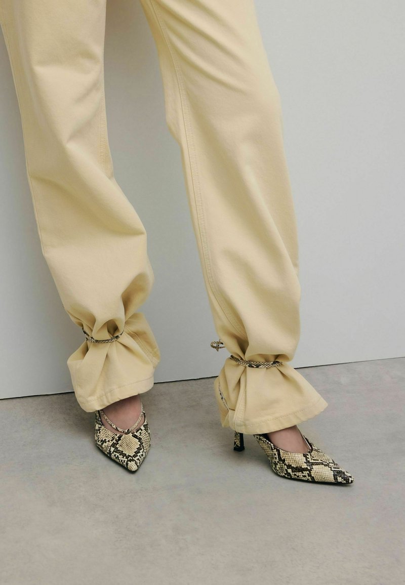 Escarpins à bout pointu avec un imprimé serpent noir et beige. Pantalon beige à cordon de serrage à la cheville avec une coupe décontractée.