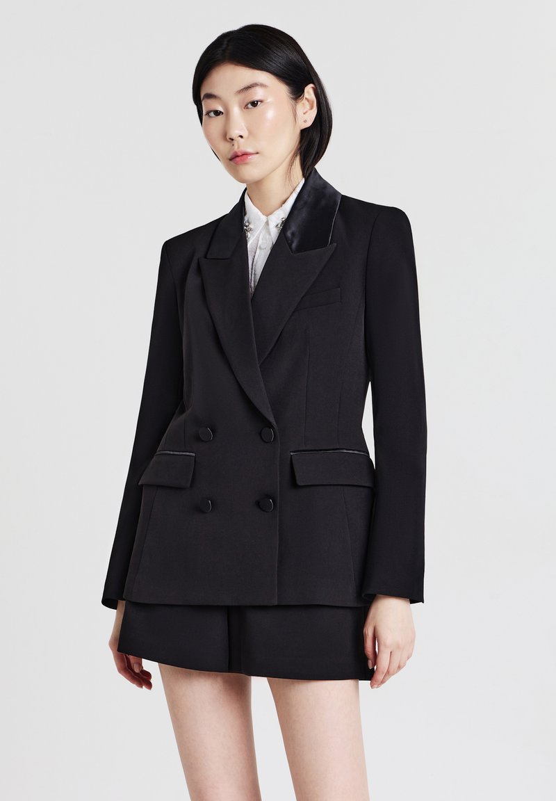Donna in un blazer doppiopetto nero con revers in satin e pantaloncini neri abbinati, in piedi davanti a uno sfondo chiaro e semplice.