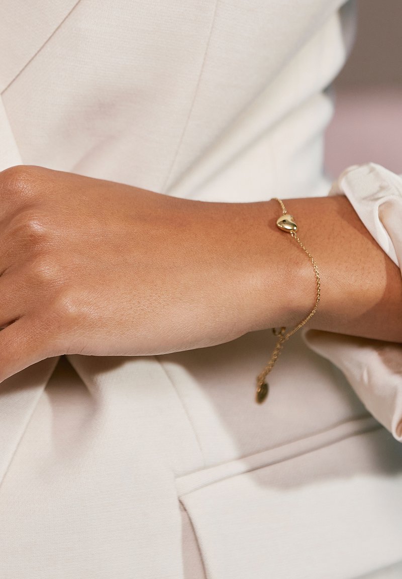 Bracciale d'oro con un ciondolo a forma di cuore e una delicata catena, indossato su un polso contro uno sfondo di tessuto chiaro.