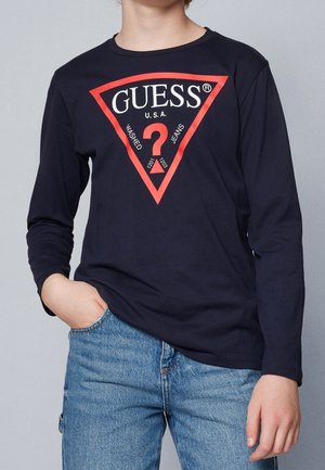 Personne portant une chemise à manches longues bleu marine avec un logo rouge en triangle Guess et un jean bleu clair, une main dans la poche, sur un fond uni.