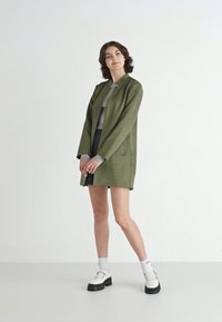 Manteau vert olive en suède, de coupe droite, avec poches latérales et manches longues, associé à un haut gris et une jupe plissée.