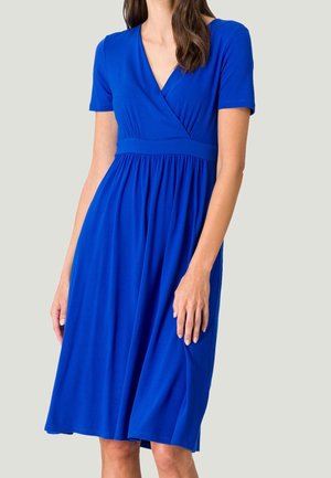 Femme portant une robe bleu roi mi-longue avec des manches courtes et un corsage croisé en V, debout devant un fond clair uni.