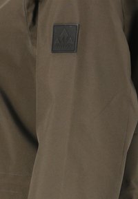 Brauner Stoff mit einem schwarzen quadratischen Aufnäher, der drei Berggipfel zeigt und darunter das Wort "WHISTLER" aufgestickt ist.