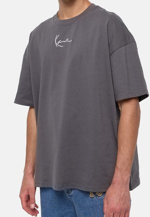 Persona que lleva una camiseta suelta de color gris oscuro con mangas cortas y texto en cursiva blanca en el pecho, combinada con jeans azules.