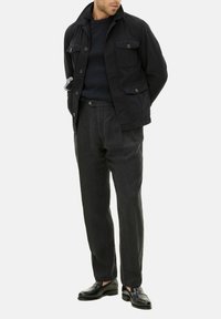 Giacca blu scuro con colletto e molteplici tasche, abbinata a un maglione blu strutturato e pantaloni antracite. Scarpe in pelle nera completano l'outfit.