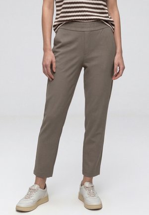 Personne portant un pantalon slim taupe avec une taille élastique et des baskets blanches, debout devant un fond clair uni.