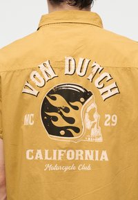 Tył musztardowo-żółtej koszuli z vintage czaszką noszącą czarny kask z płomieniami i tekstem "Von Dutch 29 MC California Motorcycle Club."