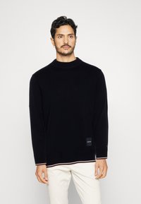 Uomo con capelli ricci scuri che indossa un maglione nero a maniche lunghe e pantaloni bianchi, in piedi contro uno sfondo chiaro e uniforme.