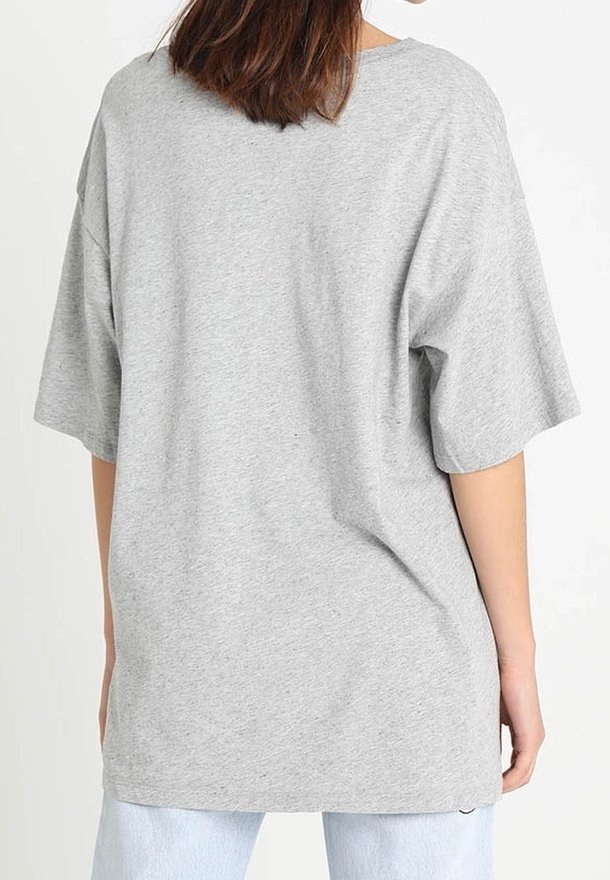 Femme aux cheveux bruns mi-longs portant un t-shirt ample gris clair à manches courtes et un jean bleu clair, vue de dos.