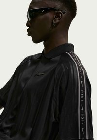 Άτομο που φοράει μαύρο πόλο της Nike με το λογότυπο στο στήθος και ρίγα με το εμπορικό σήμα κατά μήκος του μανικιού, επίσης φοράει μαύρα γυαλιά ηλίου και ένα μικρό σκουλαρίκι κρίκο.