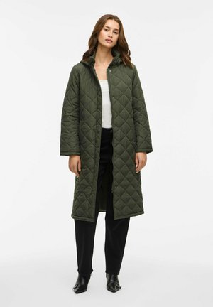 Femme portant un long manteau matelassé vert foncé sur un haut blanc et un pantalon noir, se tenant contre un fond blanc uni.