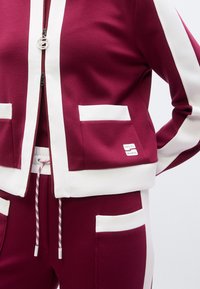 Μακρυμάνικο αθλητικό μπουφάν σε μπορντό χρώμα  με λευκές ρίγες και τσέπες, που διαθέτει φερμουάρ μπροστά και κορδόνι στη μέση. Μαλακό ύφασμα με ετικέτα λογότυπου.