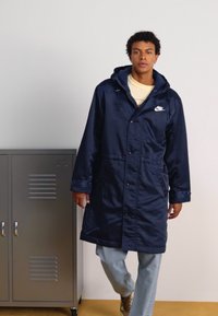 Giovane uomo che indossa un lungo cappotto Nike blu navy con cappuccio, jeans chiari e sneakers beige, in piedi accanto a un armadietto metallico grigio.