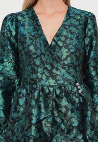 Wrap dress in a dark fabric featuring metallic green floral patterns. It has long sleeves, a deep V-neck, and decorative buttons at the waist.