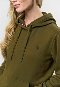 Olive zöld kapucnis pulóver, puha anyagból készült, fém végekkel ellátott húzózsinórral és egy kis fekete logóval az elején.