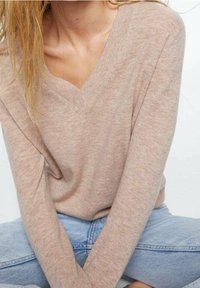 Beiger Strickpullover mit V-Ausschnitt und langen Ärmeln, mit weicher Textur. Kombiniert mit hellblauen Jeans.