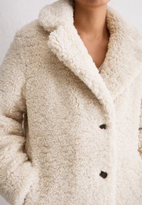 Boucle shearling jasje in crème, met een geknipt kraagje, knoopsluiting en textielstof. Zachte, fuzzy buitenkant met twee zijzakken.
