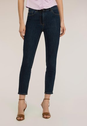 Mujer que lleva jeans ajustados azul oscuro combinados con sandalias de tacón alto en color marrón claro y una blusa rosa claro fruncida.