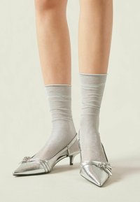 Tacones de charol plateado con punta afilada y tacón de gatito, adornados con un lazo decorativo, combinados con calcetas de tobillo plateadas y brillantes.