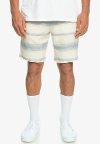 Pánské šortky s béžovými a světlemodrými horizontálními pruhy. Tkanina má texturovaný pocit, zdobena bílým prošíváním. Bílé ponožky a tenisky jsou součástí.