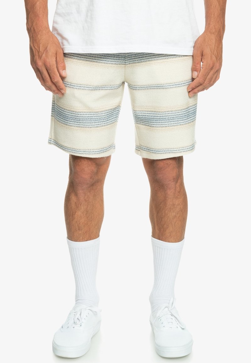 Pánské šortky s béžovými a světlemodrými horizontálními pruhy. Tkanina má texturovaný pocit, zdobena bílým prošíváním. Bílé ponožky a tenisky jsou součástí.