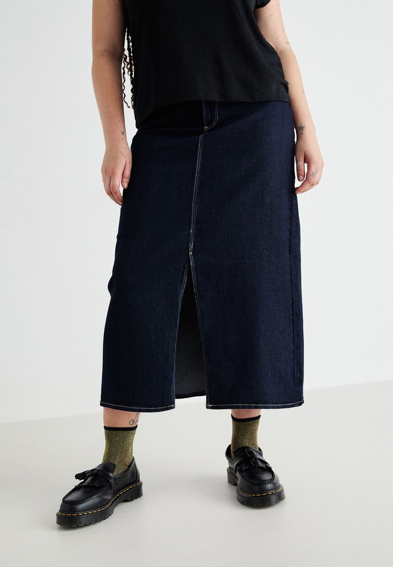 Gonna maxi in denim blu scuro con spacco frontale, caratterizzata da cuciture a contrasto e vita aderente. Abbinata a scarpe nere e calzini verdi.